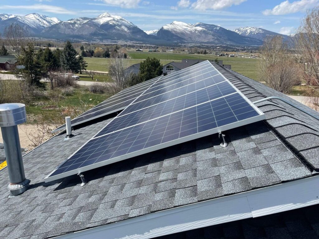 Solar panels mounted on a shingled rooftop with a scenic background of snow-capped mountains, green fields, and scattered trees under a partly cloudy sky.