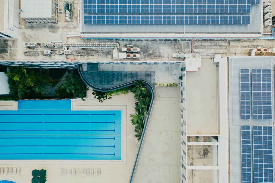 Aerial view of a building complex with a large rectangular swimming pool, a smaller pool, solar panels on rooftops, and surrounding greenery. The layout is geometric and modern.