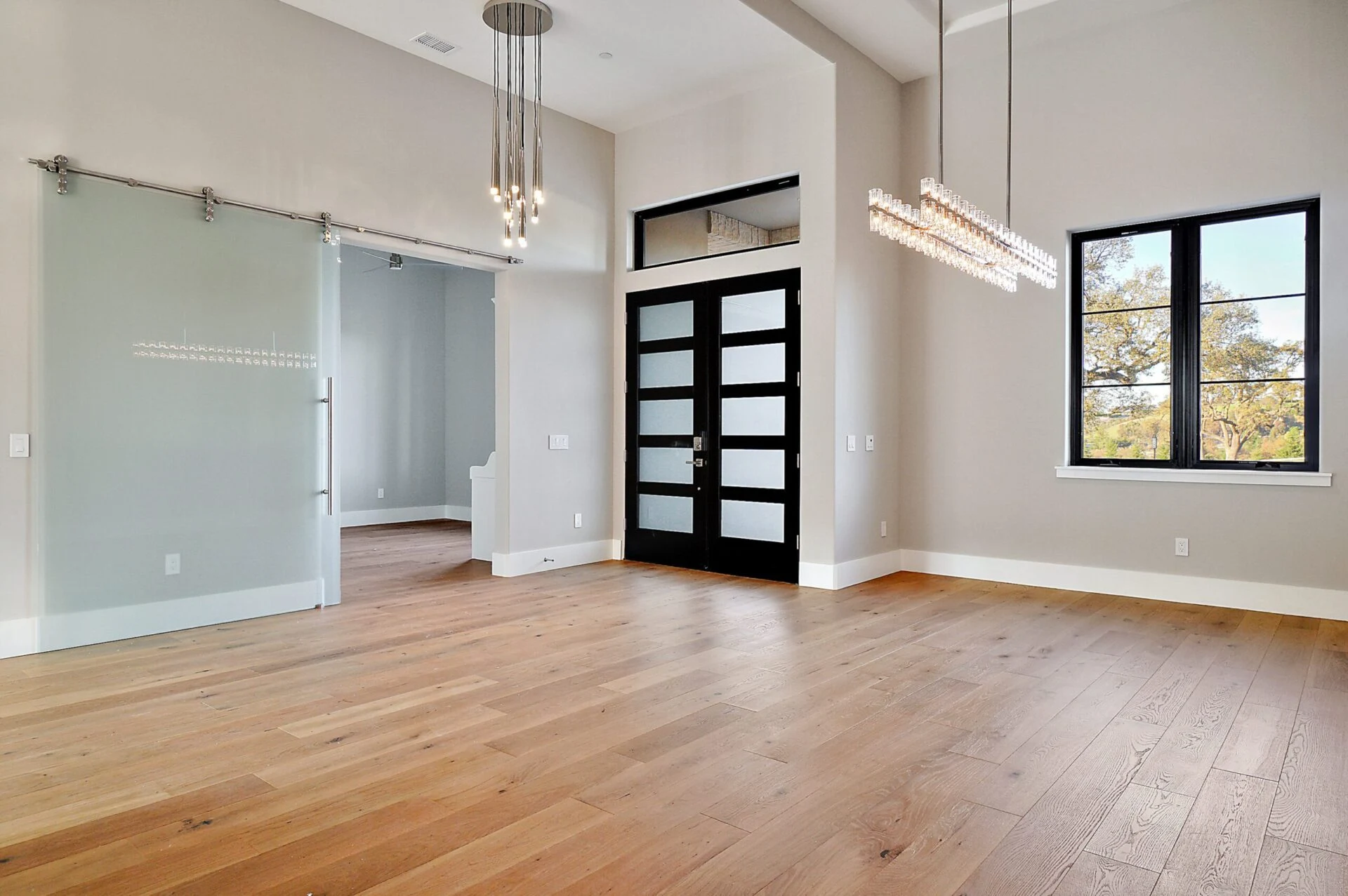 Spacious modern room with light wood flooring, high ceilings, two contemporary chandeliers, a black-framed glass front door, a large window, and a frosted glass sliding door leading to another area.