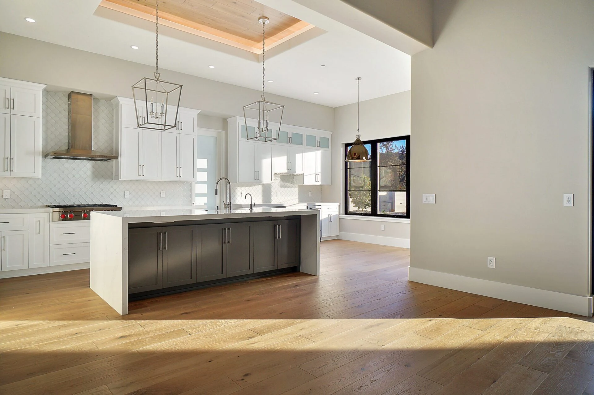 Modern kitchen with white cabinets, a large dark island, pendant lights, stainless steel appliances, light wood flooring, and large windows letting in natural light.