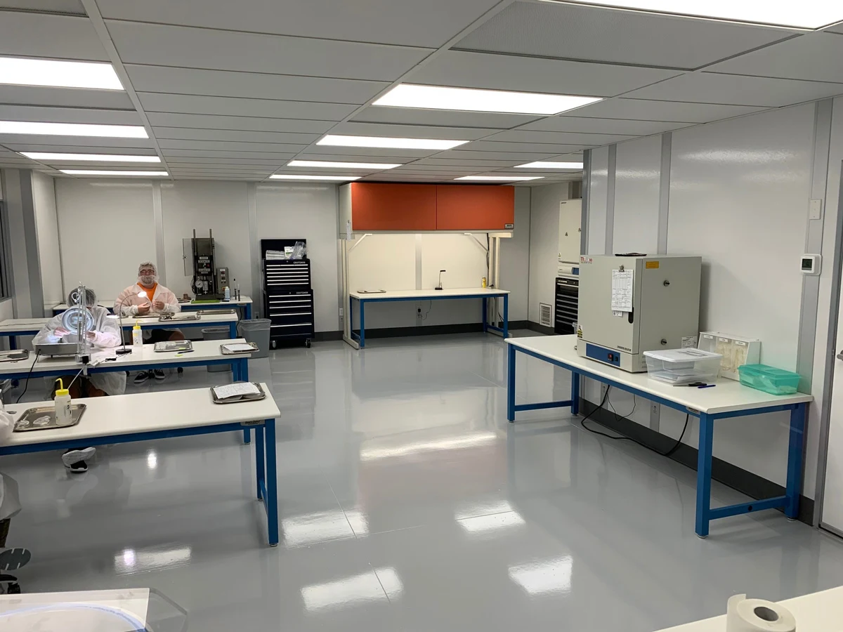 A clean, modern laboratory with white walls and floors, several workbenches, scientific equipment, and one person in protective gear seated at a table on the left side of the room.