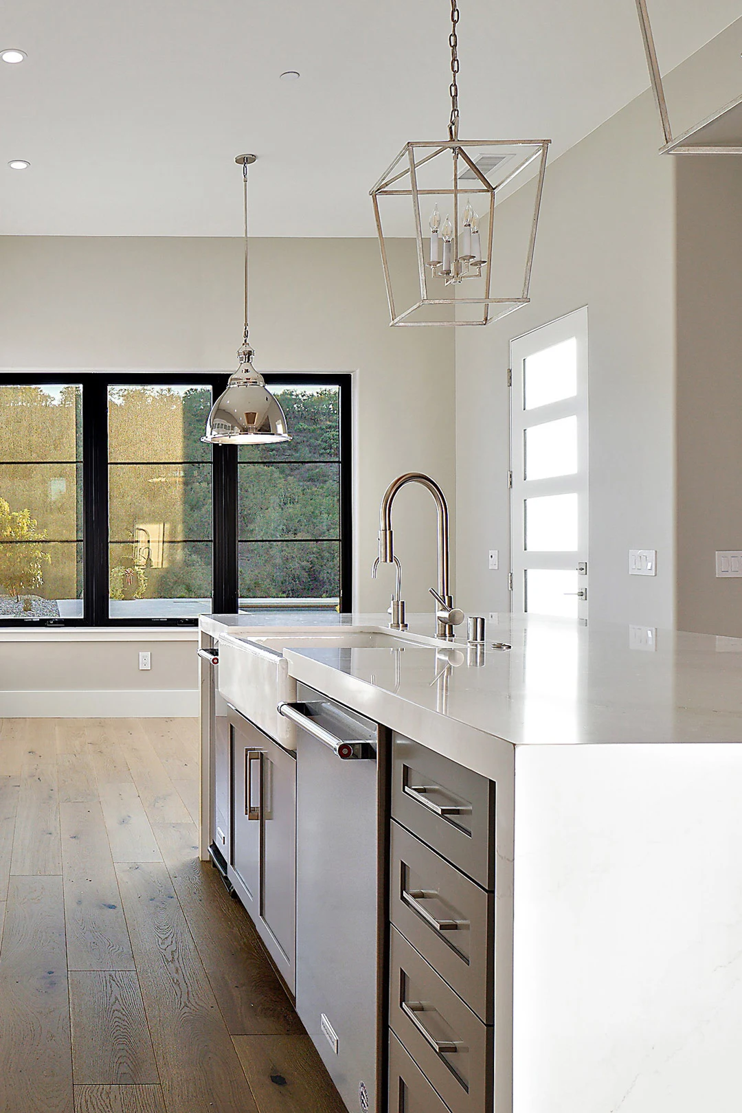Modern kitchen with a white island countertop, stainless steel appliances, light wood flooring, large black-framed windows, and contemporary pendant lighting fixtures hanging from the ceiling.