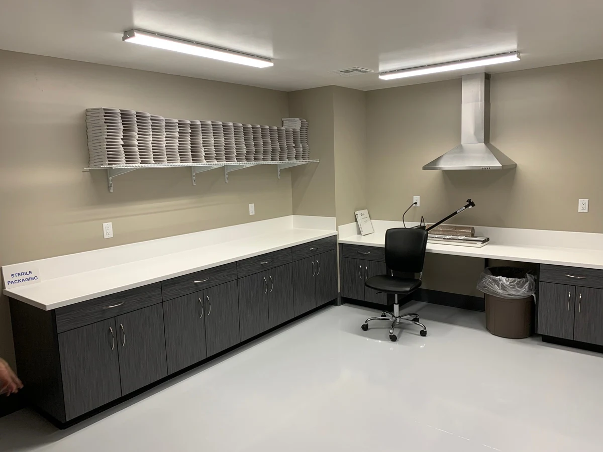 A clean, modern workroom with dark cabinetry, white countertops, a black office chair, a wall-mounted shelf with stacked trays, and a stainless steel range hood. A sign reads STERILE PACKAGING. The floor and walls are light-colored.