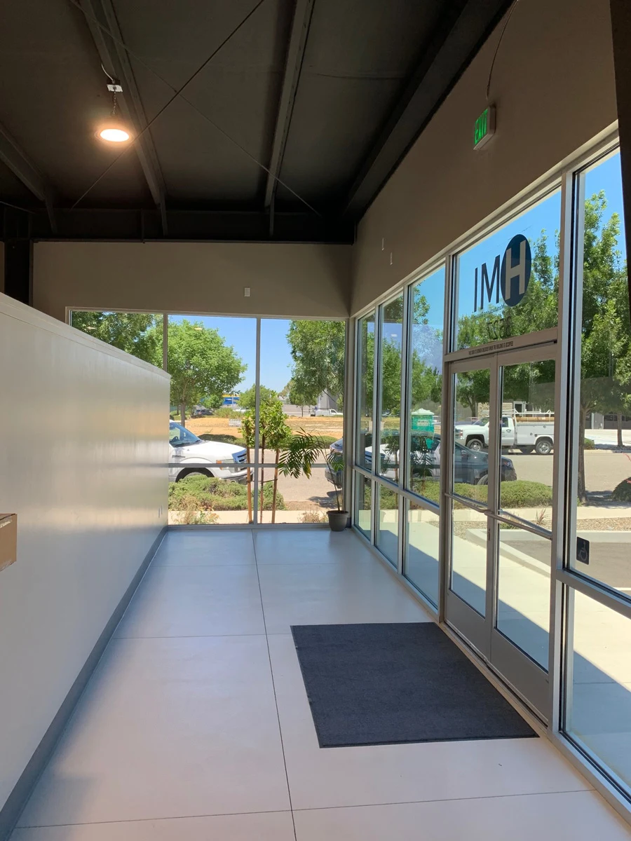 A spacious building entrance with large glass windows and doors, letting in sunlight. A dark doormat is on the floor, and trees, cars, and a sidewalk are visible outside. The letters IMH appear on the glass door.