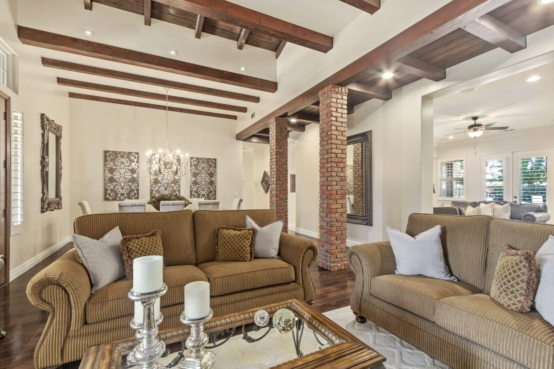 A spacious living room with exposed wooden ceiling beams, brick columns, tan sofas with cushions, a glass coffee table with candles, and a view into a dining area with a chandelier.
