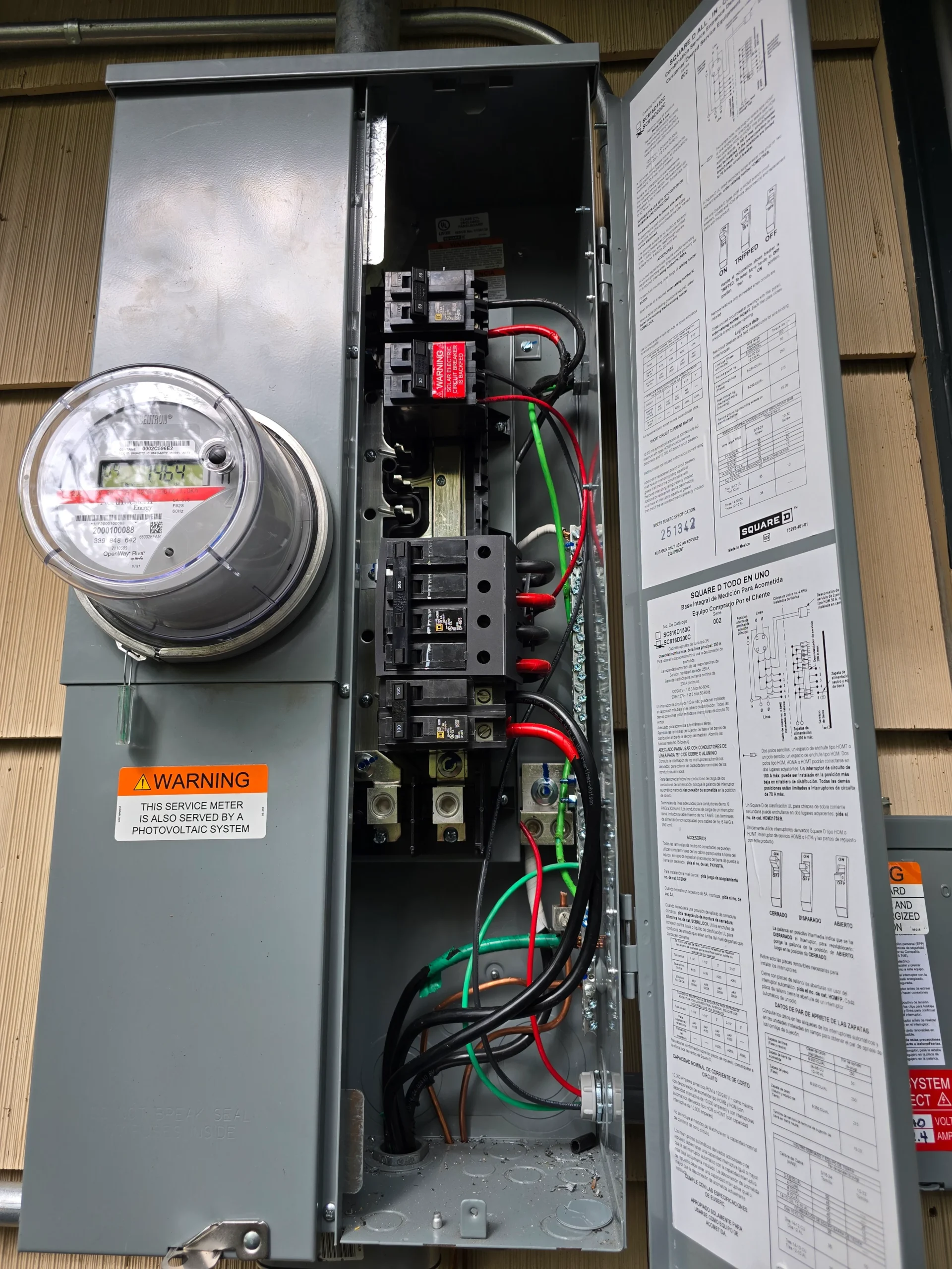 An open electrical panel with visible circuit breakers, wiring, a meter on the left, safety warning label, and wiring diagrams on the inside of the door. The panel is mounted on an exterior wall.