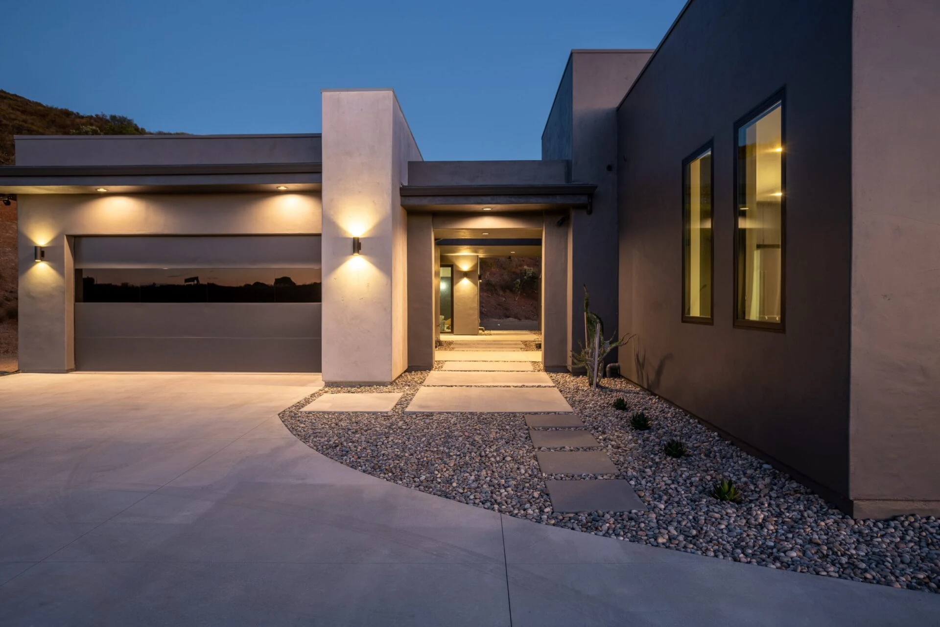 Modern single-story house at dusk with large windows, concrete walls, a flat roof, illuminated entryway, and landscaped yard featuring stone pathways and rocks.