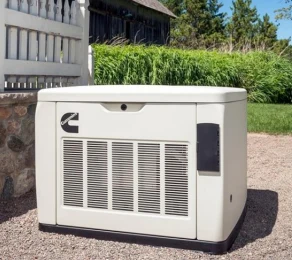 A white Cummins standby generator sits on a gravel area outside a house, with green grass and bushes in the background.
