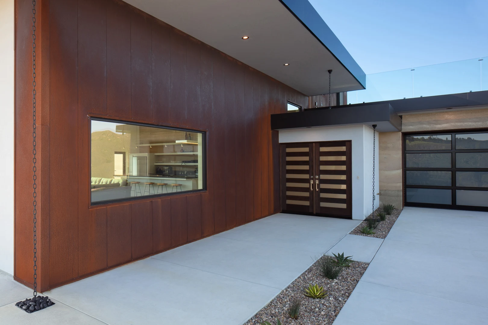 A modern house exterior with a large wood-paneled wall, horizontal window, double front doors with glass panels, a glass-paneled garage door, and minimalist landscaping with rocks and succulents.