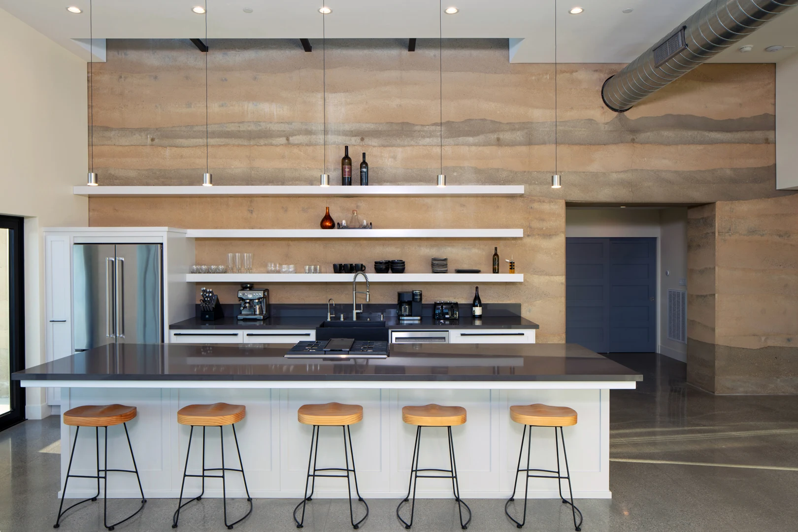 Modern kitchen with a large island, five wooden stools, open shelves with bottles and glasses, stainless steel appliances, and a textured tan and beige feature wall. Industrial lighting and exposed ductwork overhead.