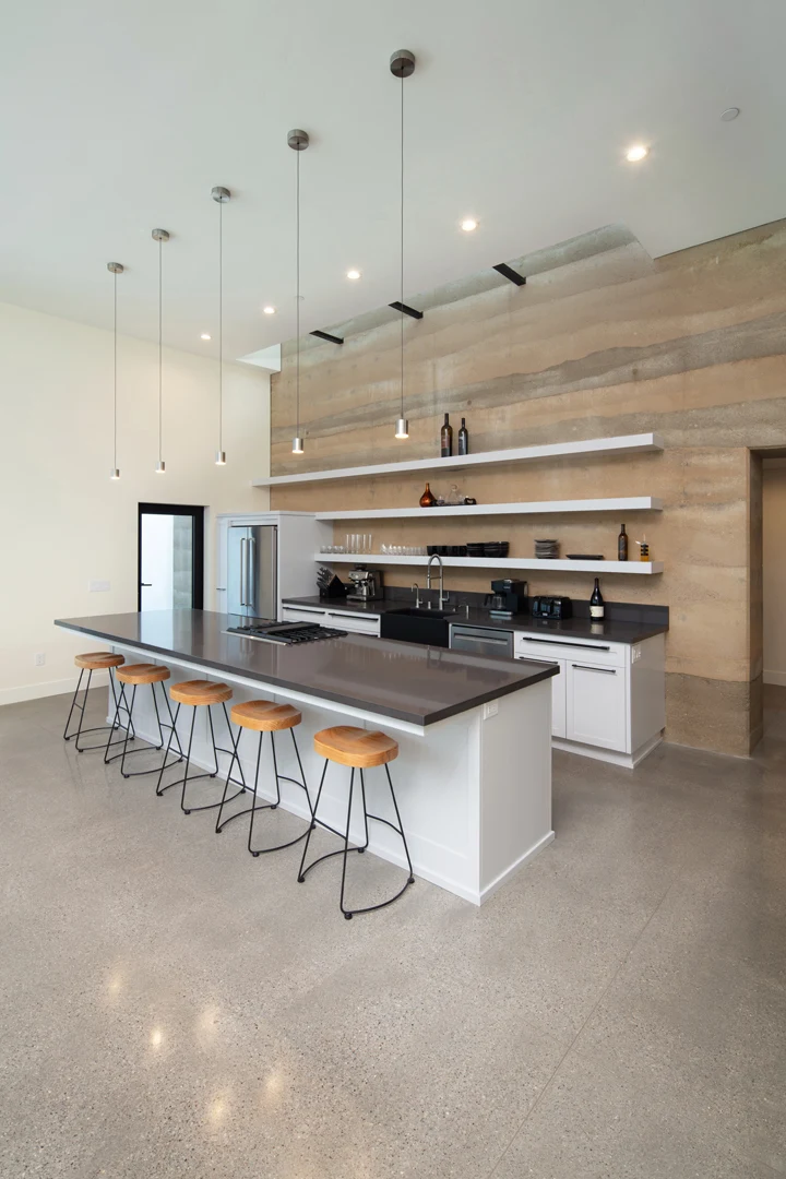 Modern kitchen with a large island, black countertop, four wooden bar stools, stainless steel appliances, floating shelves with decor, and a textured beige accent wall. The space is bright with minimalist design.
