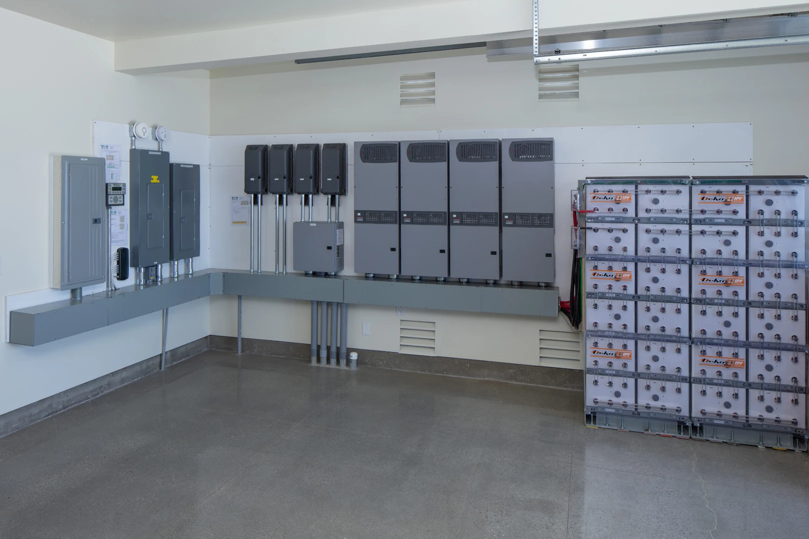 A clean, modern utility room with electrical panels and equipment mounted on the wall, and a stack of large batteries in clear cases to the right, all set against a polished concrete floor.