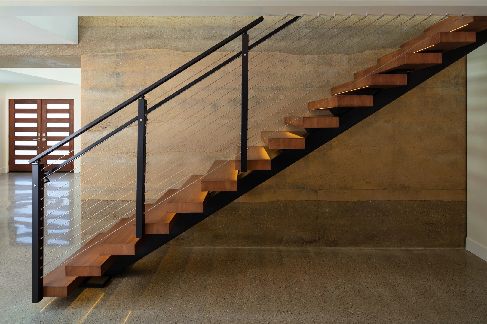 Modern wooden staircase with metal railing and cable wires, set against a textured beige wall and polished concrete floor in a contemporary home interior.