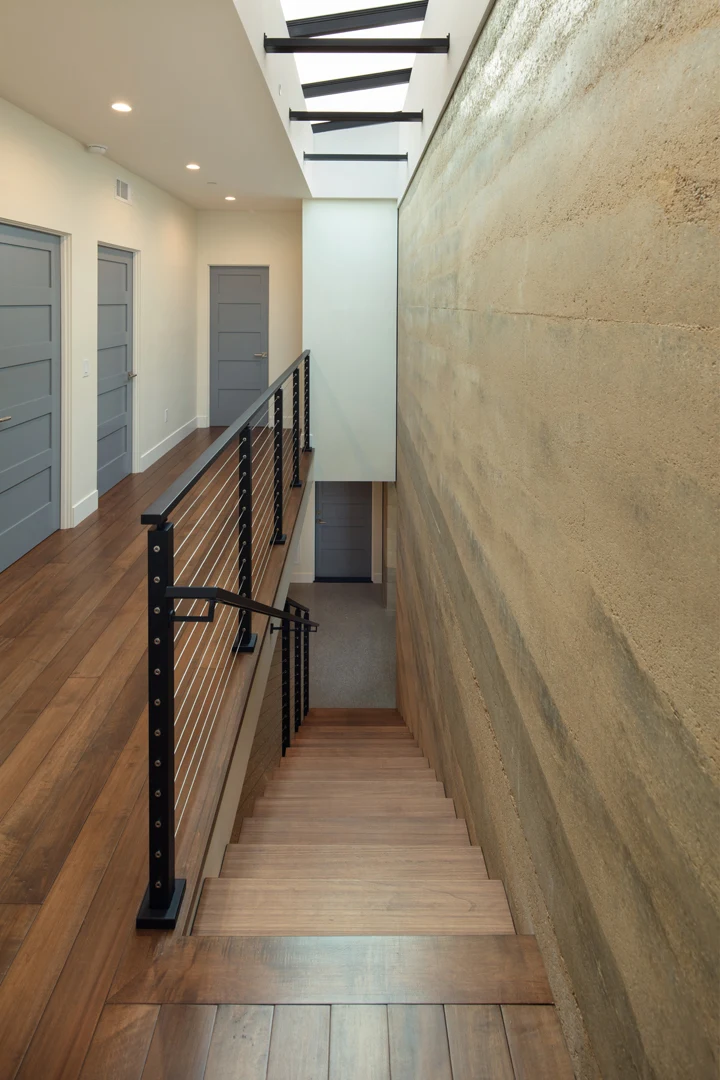 A modern hallway with wooden floors, gray doors, and a staircase with metal railings descends alongside a textured beige wall. Skylights above let in natural light.