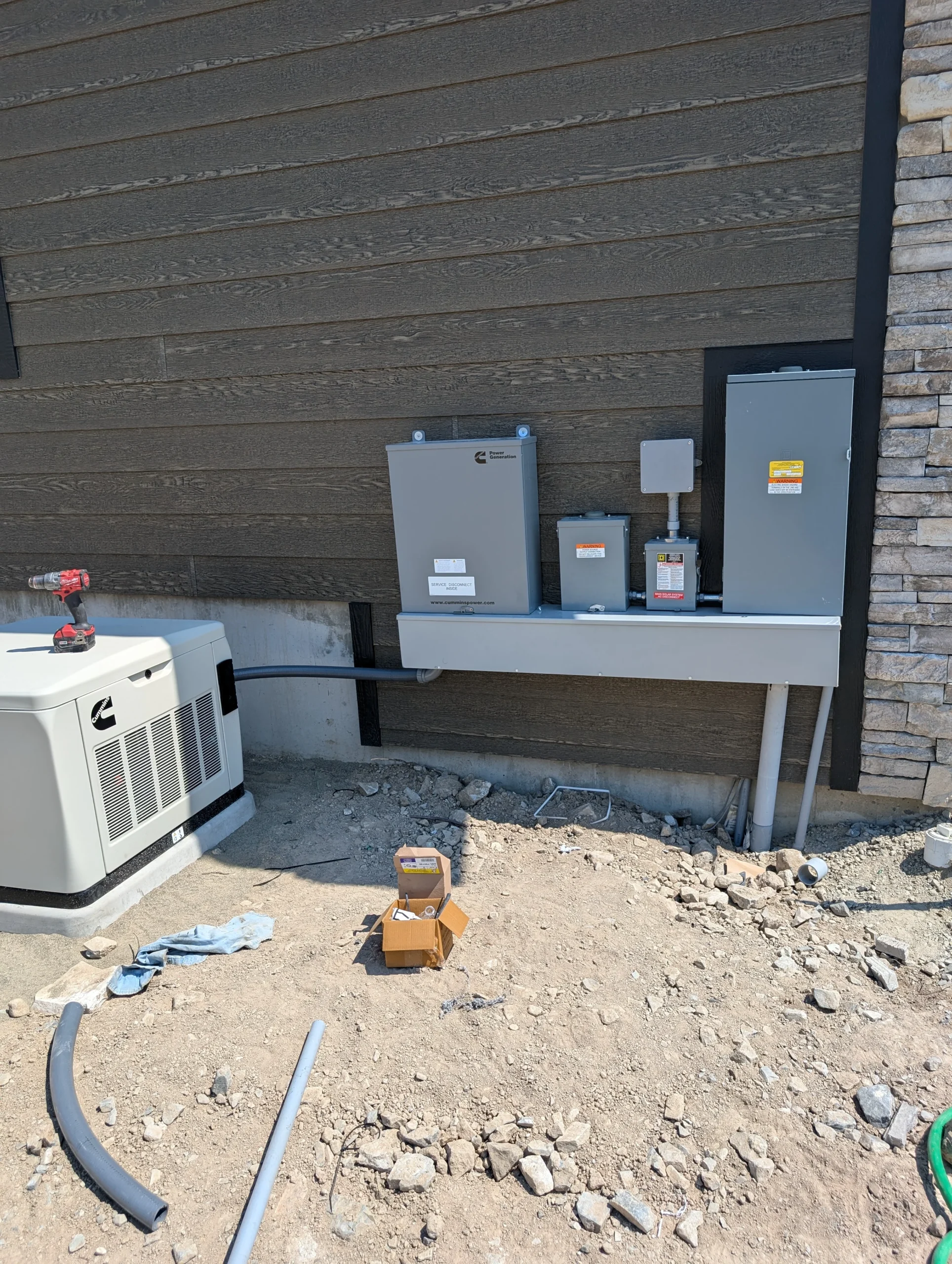 Outdoor electrical panels and a generator are mounted on the side of a building with a stone and wood exterior. Electrical conduit lines run from the generator to the panels. Tools and a small box are on the rocky ground.