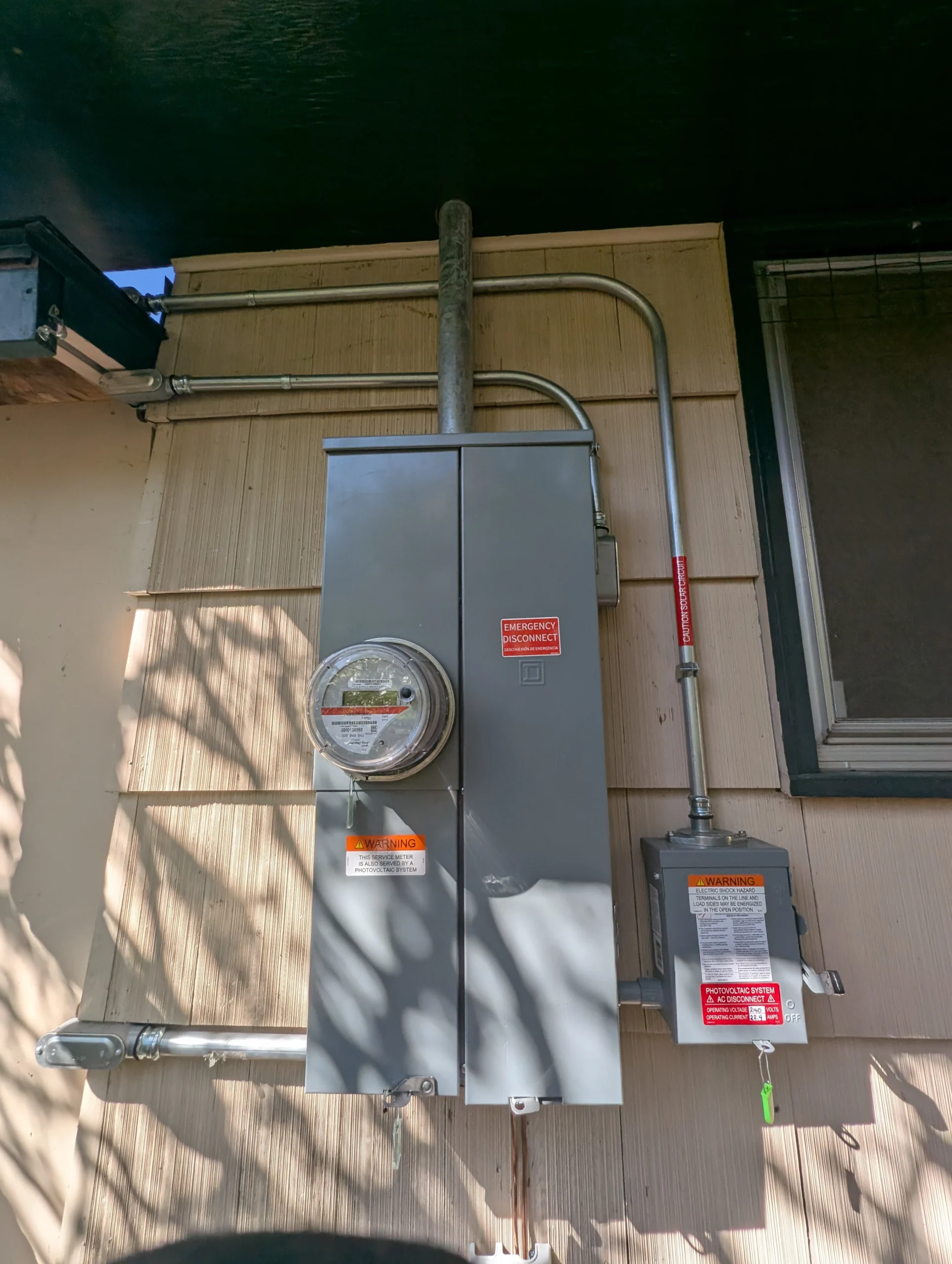 An outdoor electric meter and gray breaker panel are mounted on a beige wall, with various conduits and a smaller control box nearby. Sunlight and shadows partially cover the equipment.