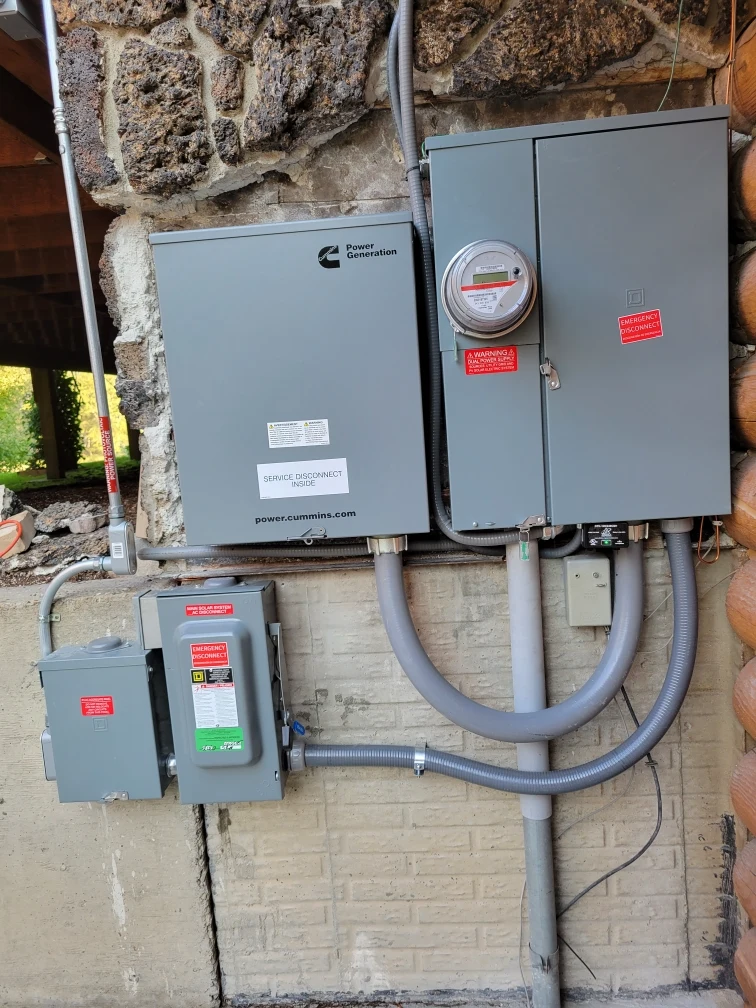Three gray electrical panels with warning labels and a circular electric meter are mounted on an exterior brick and stone wall, with several gray conduit pipes connecting the boxes.