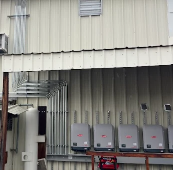 Five electrical inverter boxes are mounted on the exterior wall of a metal building. Several neatly arranged metal conduit pipes run vertically and curve along the wall above the inverters.