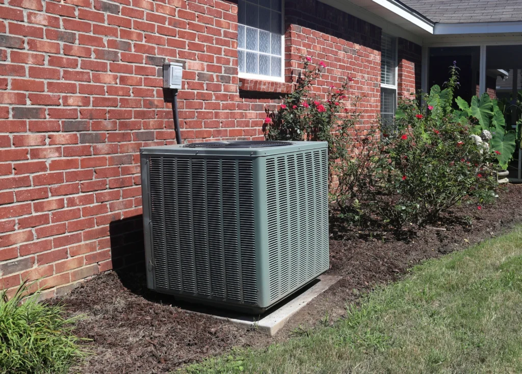 An outdoor air conditioning unit sits on a concrete slab next to a red brick house, surrounded by a garden bed with bushes and flowers, and a grassy lawn in the foreground.