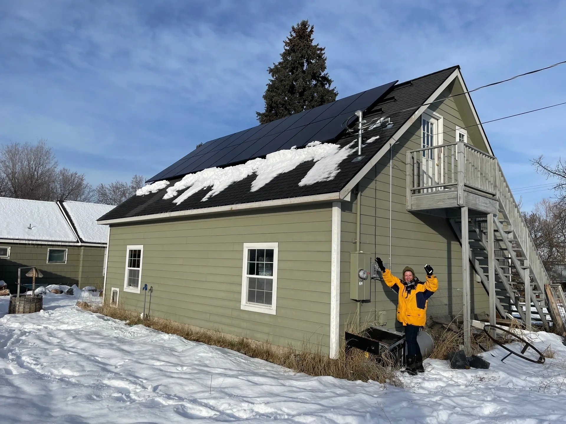 A person in a yellow jacket stands outside a green house with solar panels on the snowy roof, raising their arms in excitement. Snow covers the ground and nearby rooftops on a clear winter day.