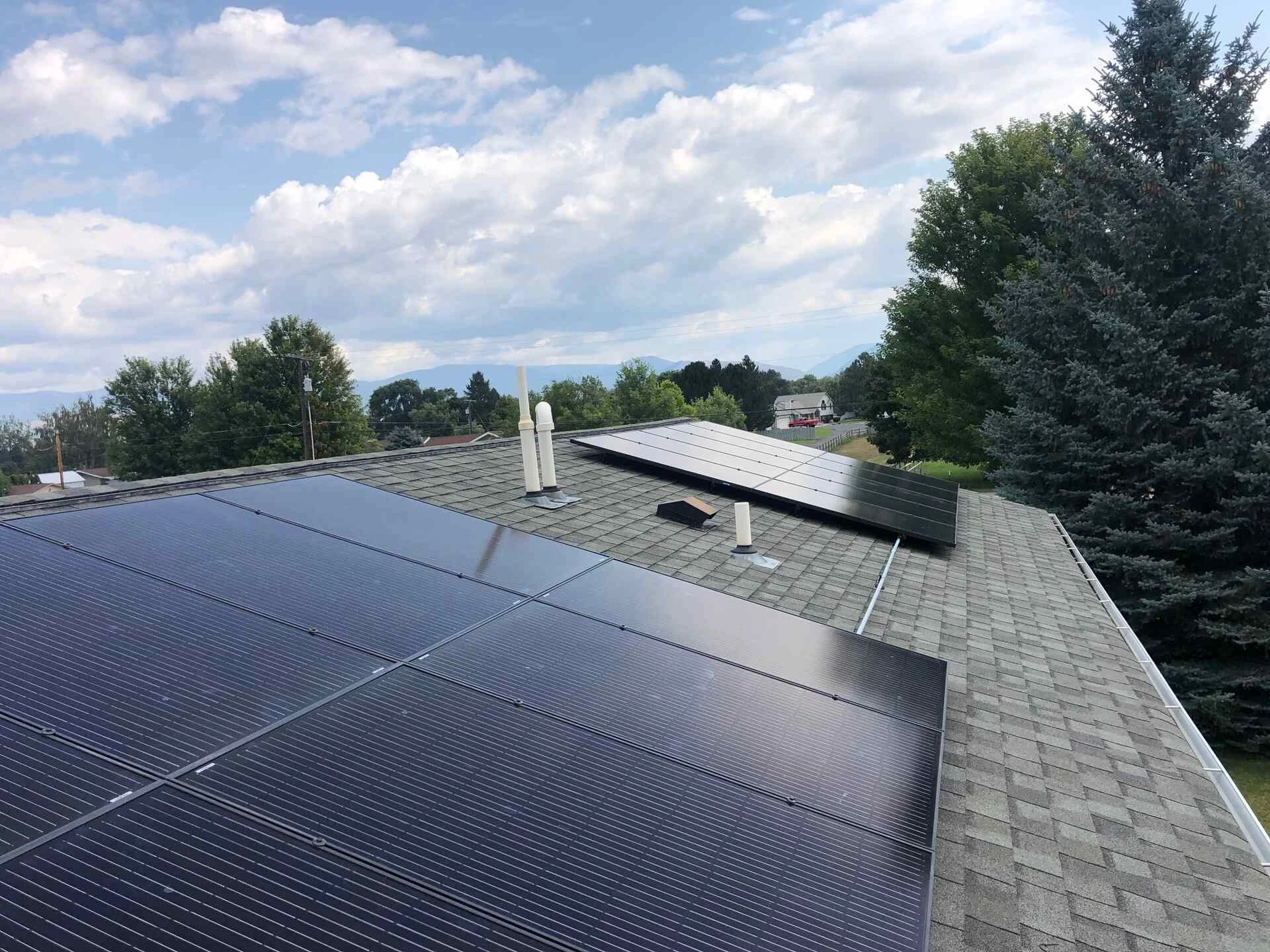 Rows of solar panels are installed on the gray shingled roof of a house, with trees and a partly cloudy sky visible in the background.