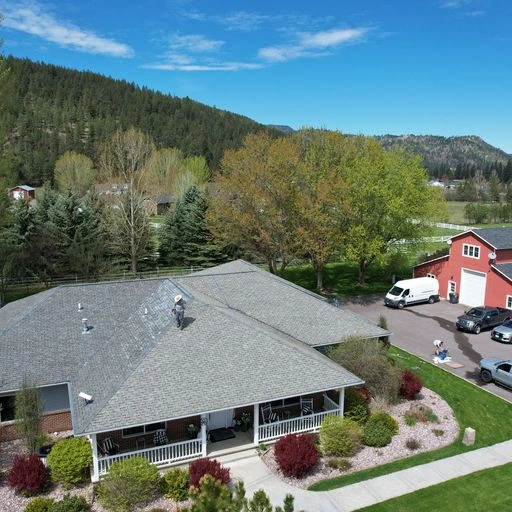 Aerial view of a house with a gray roof, front porch, and landscaped yard. Trees, a white truck, and a red and white barn are nearby, with forested hills and mountains in the background under a blue sky.