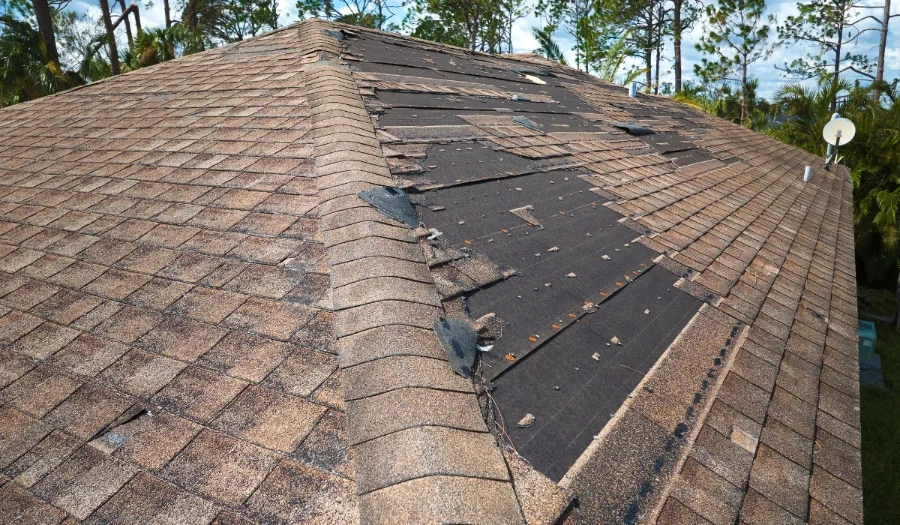 A house roof with missing and damaged shingles, exposing the black underlayment beneath. Some shingles are loose or curled, indicating storm or wind damage. Trees and a satellite dish are visible in the background.