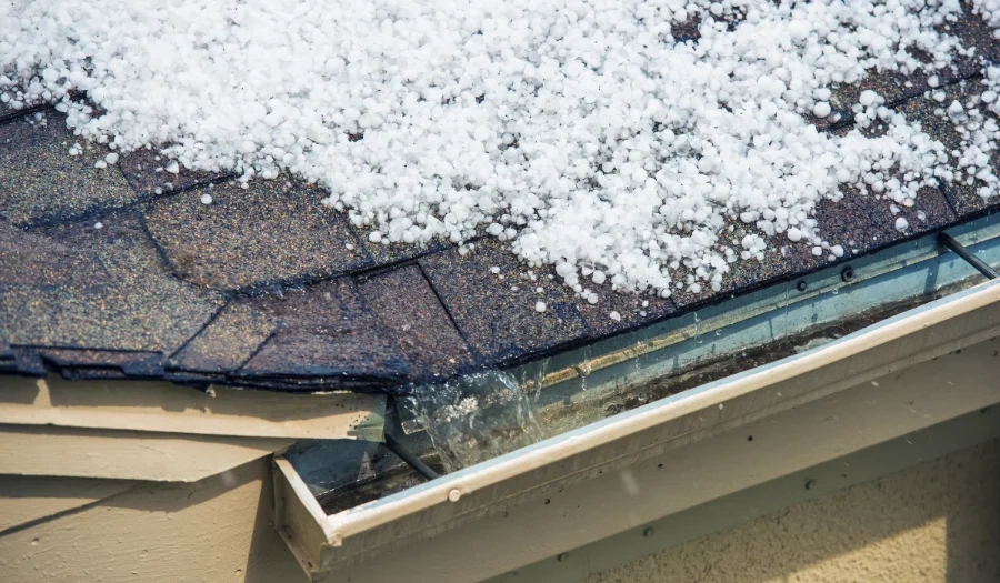 Close-up of a house roof covered with small white hailstones, with some hail collecting in the roof gutter. The roof shingles and part of the building’s exterior are visible.