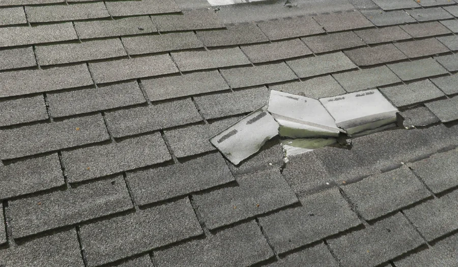 Damaged asphalt roof shingles with several shingles broken and lifted, exposing the underlying layers and creating a gap in the roof surface.