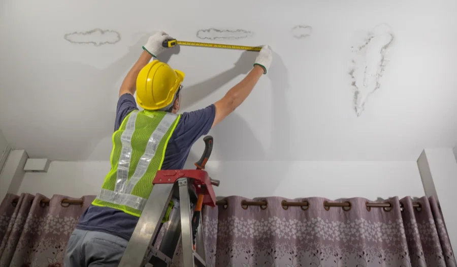A construction worker in a hard hat and safety vest stands on a ladder, measuring water-damaged spots on a white ceiling above a window with purple curtains.