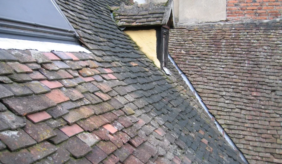 Weathered, unevenly colored clay roof tiles cover several sloped rooftops of old buildings, some with moss and darkened patches, creating a rustic and aged appearance.