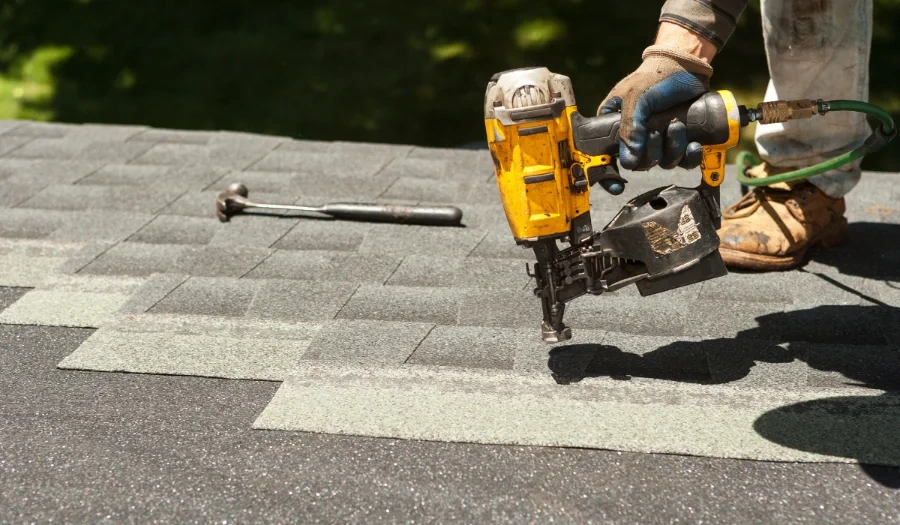 A worker uses a yellow nail gun to secure asphalt shingles on a rooftop, with a hammer lying nearby and green grass blurred in the background.