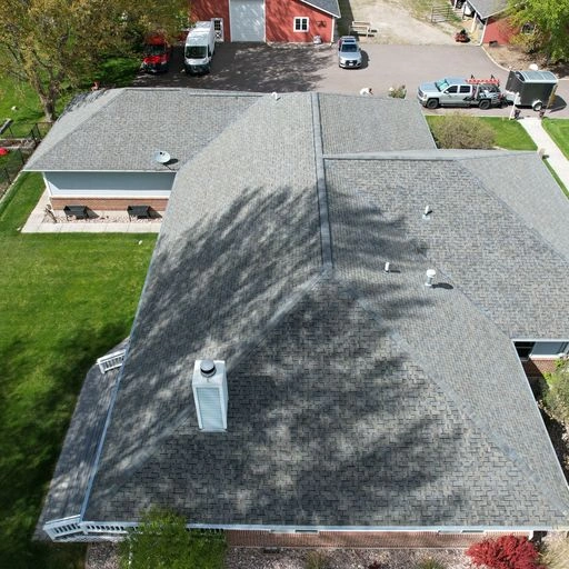 Aerial view of a house with a gray shingle roof, surrounded by green lawn and trees. Two red buildings and several vehicles are visible in the background near a driveway.