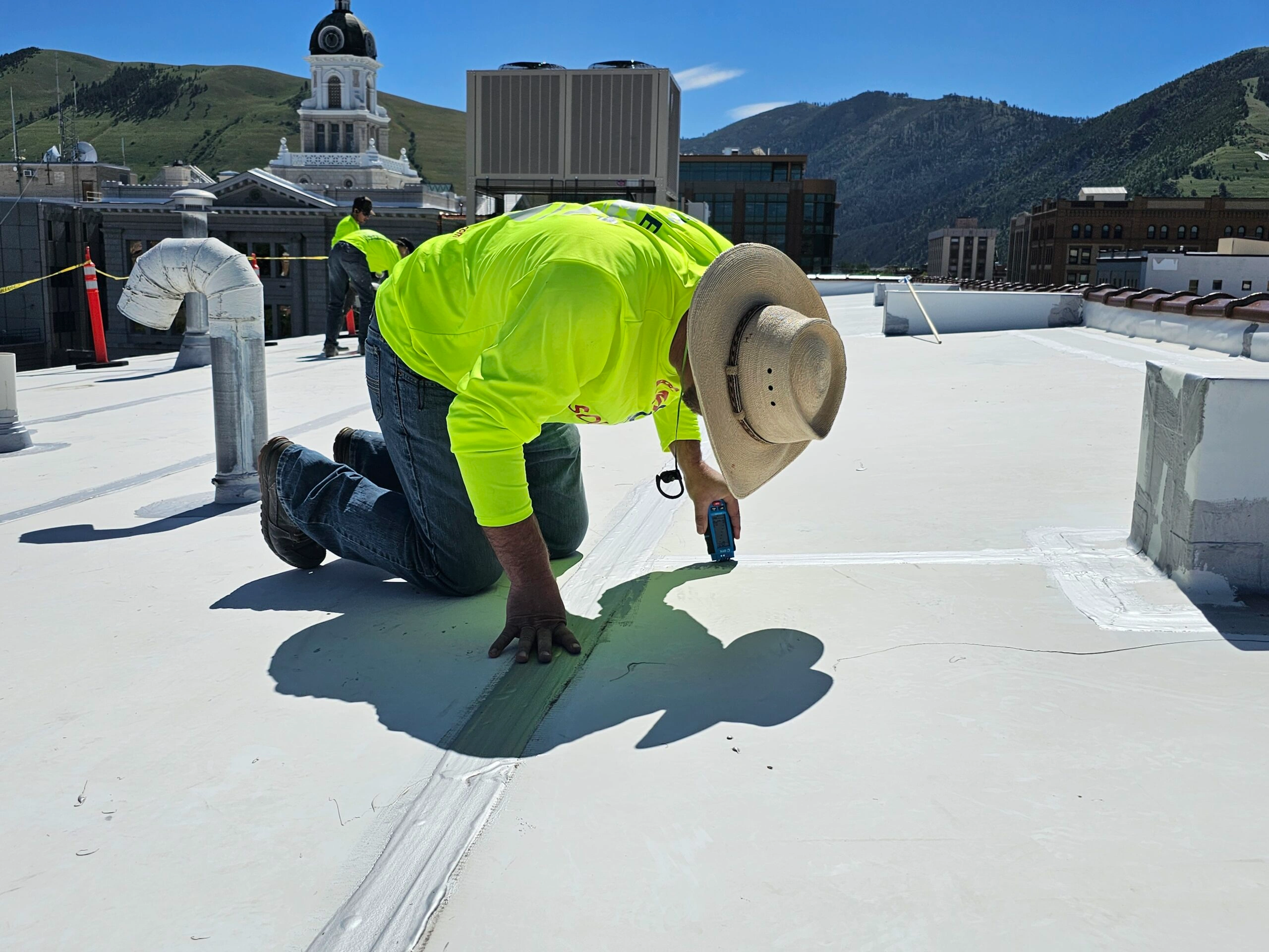 A worker in a bright yellow shirt and straw hat kneels on a rooftop, applying sealant to a seam. Hills, a courthouse dome, and other buildings are visible in the background under a sunny sky.