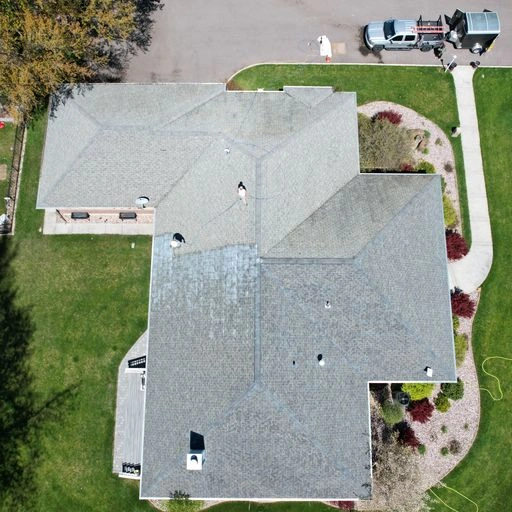 Aerial view of a house with a gray shingled roof, surrounded by green grass, landscaping with shrubs, and a curved sidewalk. A driveway with a parked truck and equipment is visible at the top.