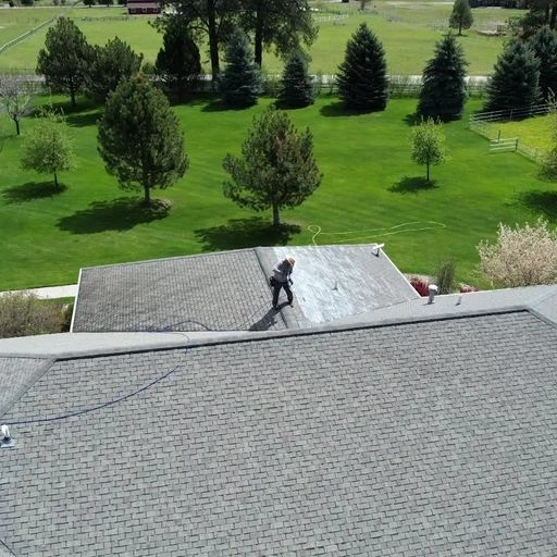 A person is seen cleaning or inspecting a gray shingled rooftop, surrounded by green lawns with trees and a road in the background. The scene is captured from above.