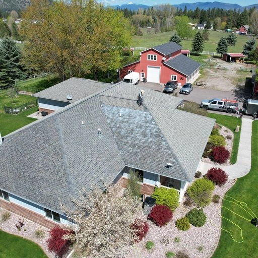 A man is working on the roof of a house surrounded by a landscaped yard with shrubs and trees. In the background, there is a red barn, vehicles, and a scenic view of mountains and fields.