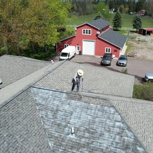 A person in a hat power-washes a gray shingled roof. In the background are a red barn, a white van, green trees, and a field. Some roof shingles appear lighter from recent cleaning.