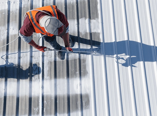 A worker wearing a safety vest and cap uses a paint roller to paint a corrugated metal roof white. The image is taken from above, showing the worker’s shadow on the roof.