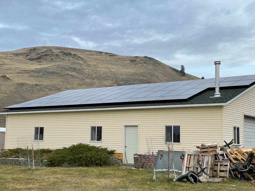 A beige building with solar panels on its roof sits in front of a grassy hill, with scattered wood, tires, and shrubs in the foreground under a cloudy sky.