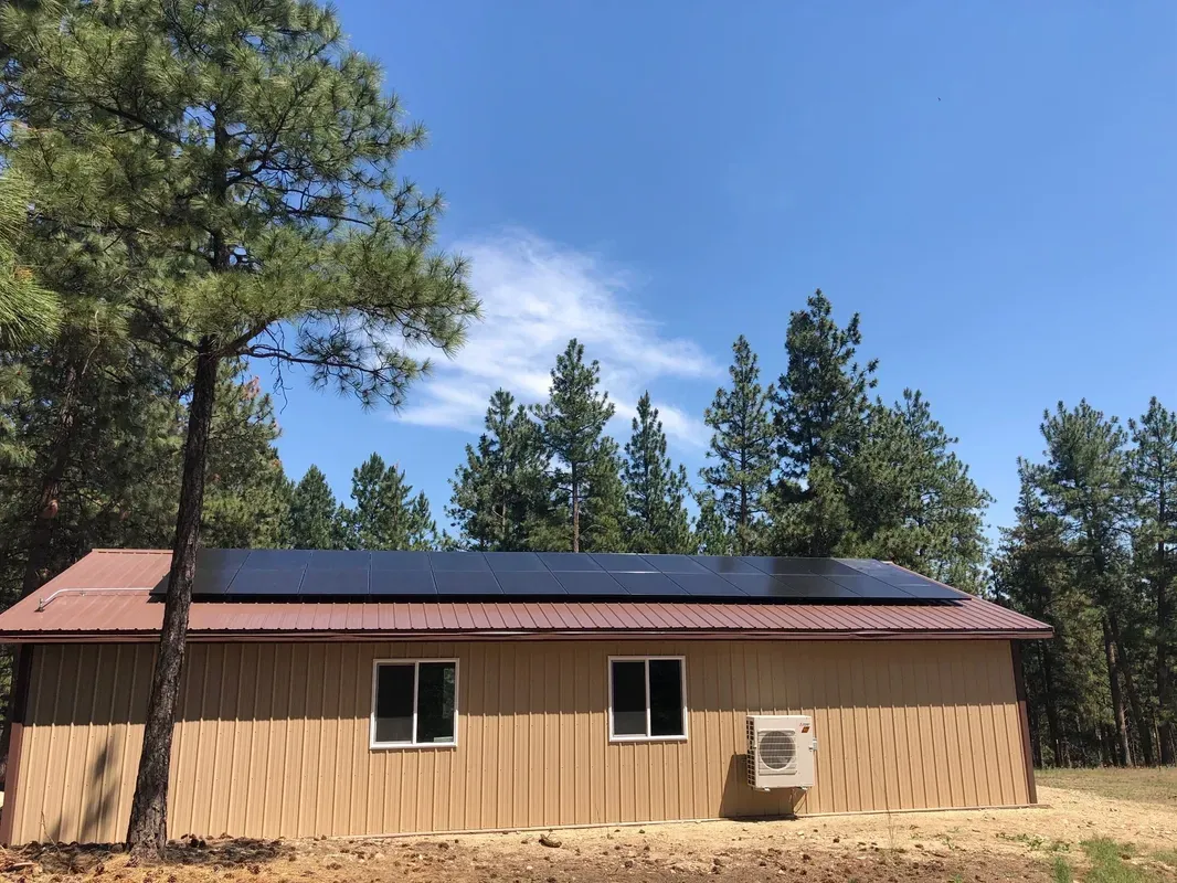A tan building with a red roof has solar panels installed on top. There are two windows and an air conditioning unit on the side. Pine trees and a clear blue sky are in the background.