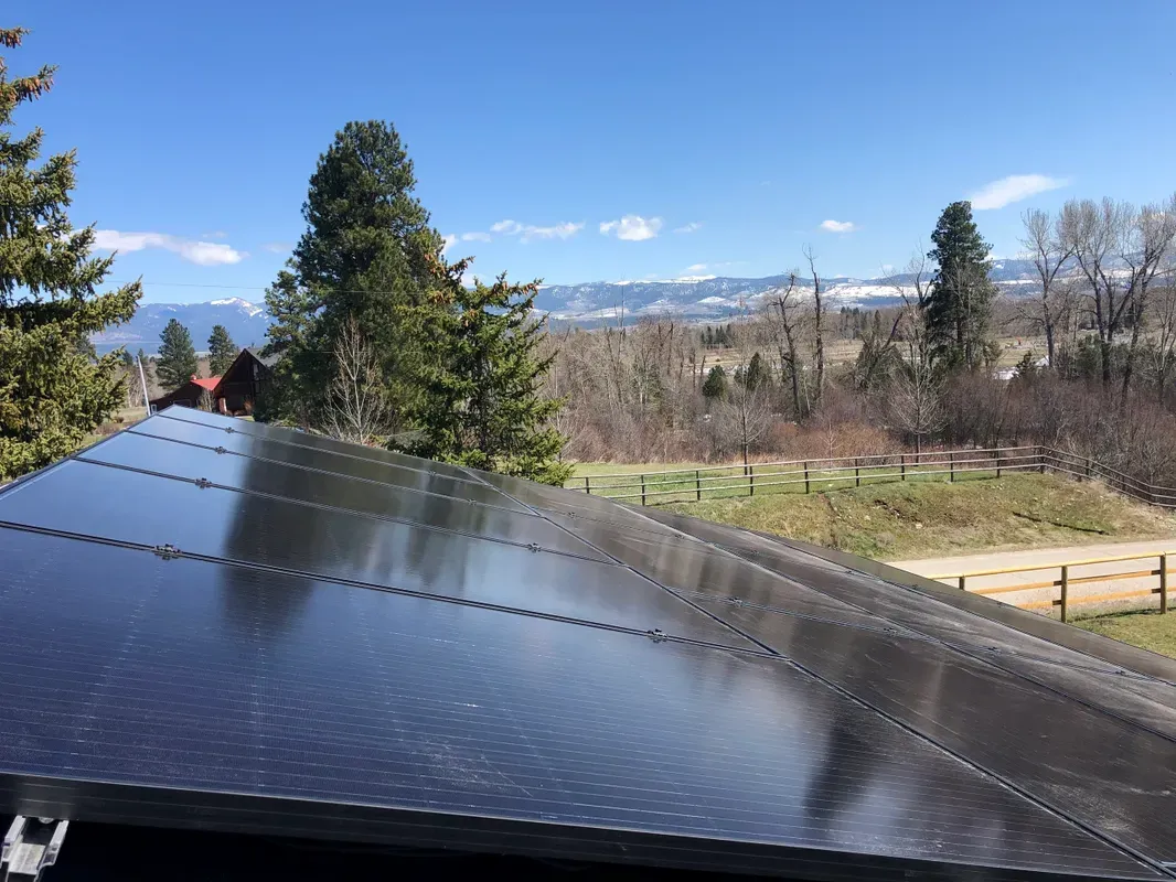 Rows of solar panels on a rooftop reflect sunlight, with trees, grass, wooden fences, and distant mountains under a blue sky in the background.
