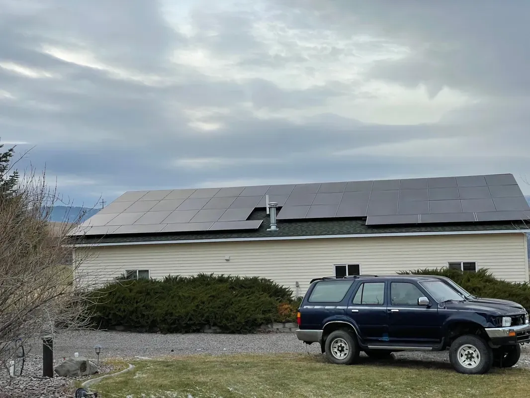 A dark SUV is parked on grass in front of a single-story house with solar panels covering its roof. The sky is cloudy, and there are bushes along the house and gravel next to the lawn.