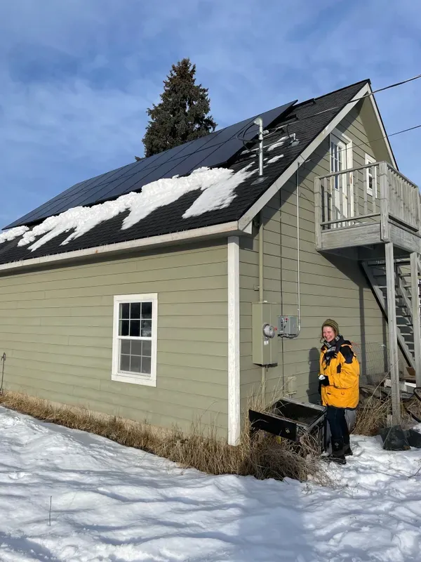A person in a yellow coat stands beside a green house with solar panels on the roof. Snow partially covers the roof, and the ground around the house is snowy. A metal staircase leads to an upper door.