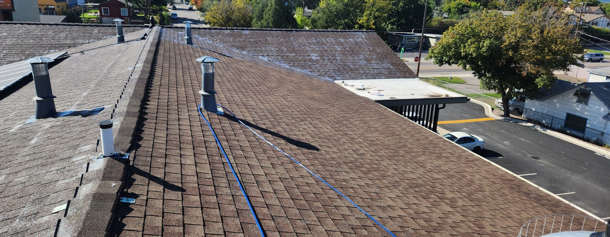 View from a rooftop showing brown shingles, several vent pipes, and blue chalk lines. In the background, trees, buildings, and parked cars are visible under a clear sky.