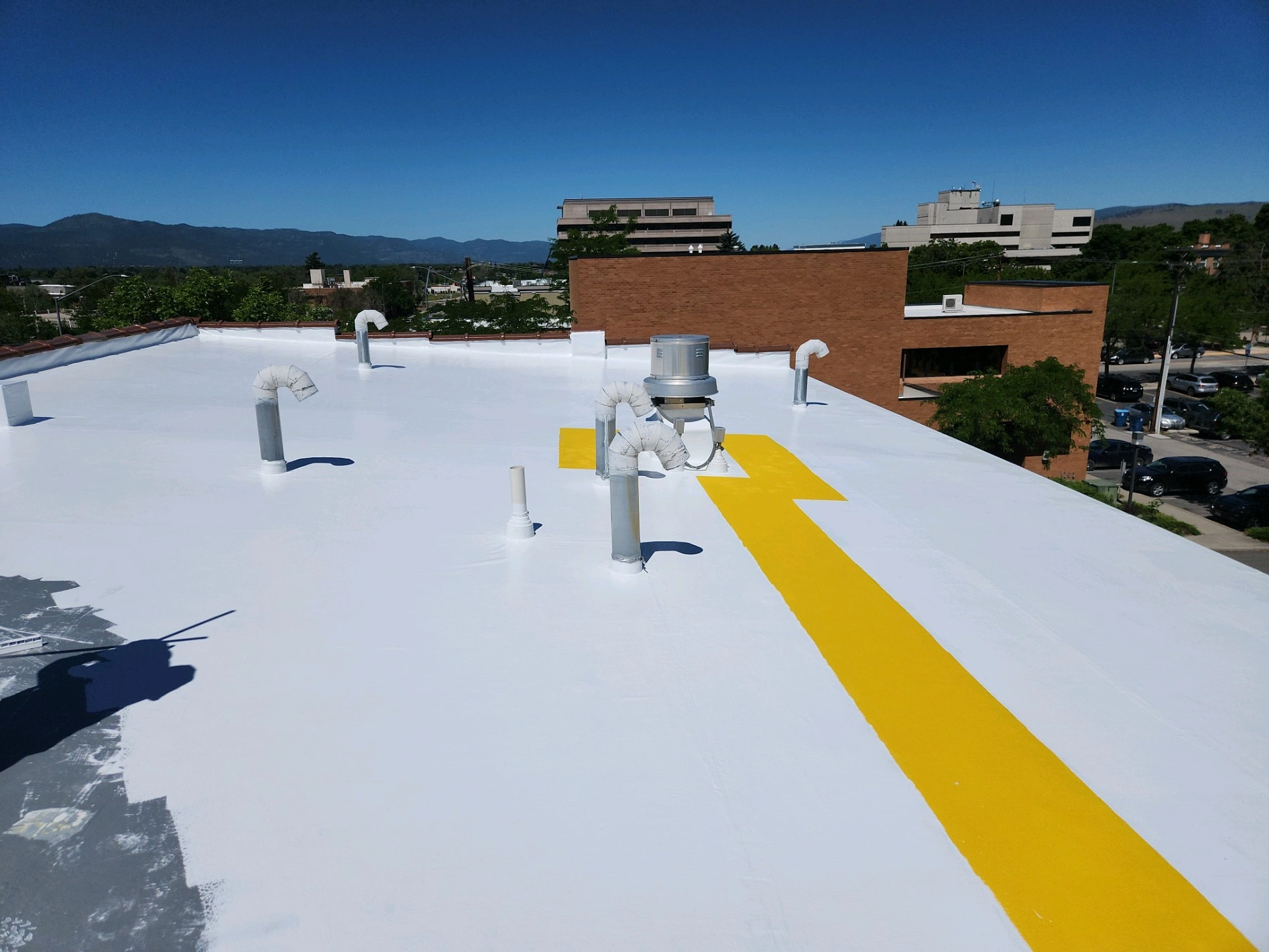 A flat white rooftop with several vent pipes and a yellow safety walkway line, set against a background of trees, buildings, and distant mountains under a clear blue sky.