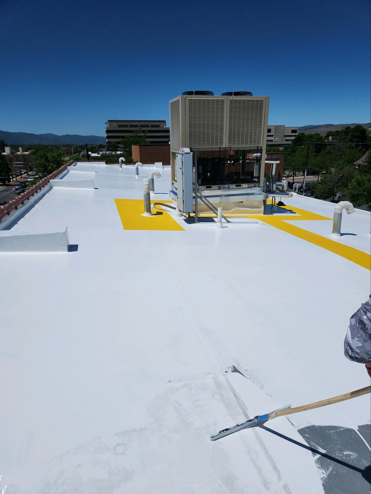 A white commercial rooftop with yellow safety markings, HVAC units, pipes, and a person using a roller to apply coating under a clear blue sky. Buildings and greenery are visible in the background.
