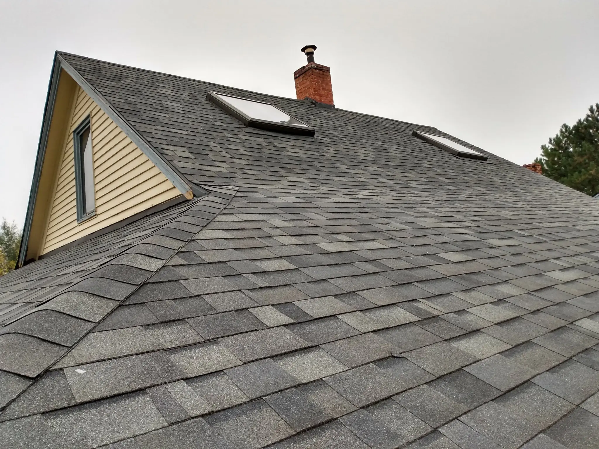 A close-up view of a house roof with gray asphalt shingles, two skylights, a brick chimney, and cream-colored siding on the triangular gable end. Trees and a cloudy sky are visible in the background.