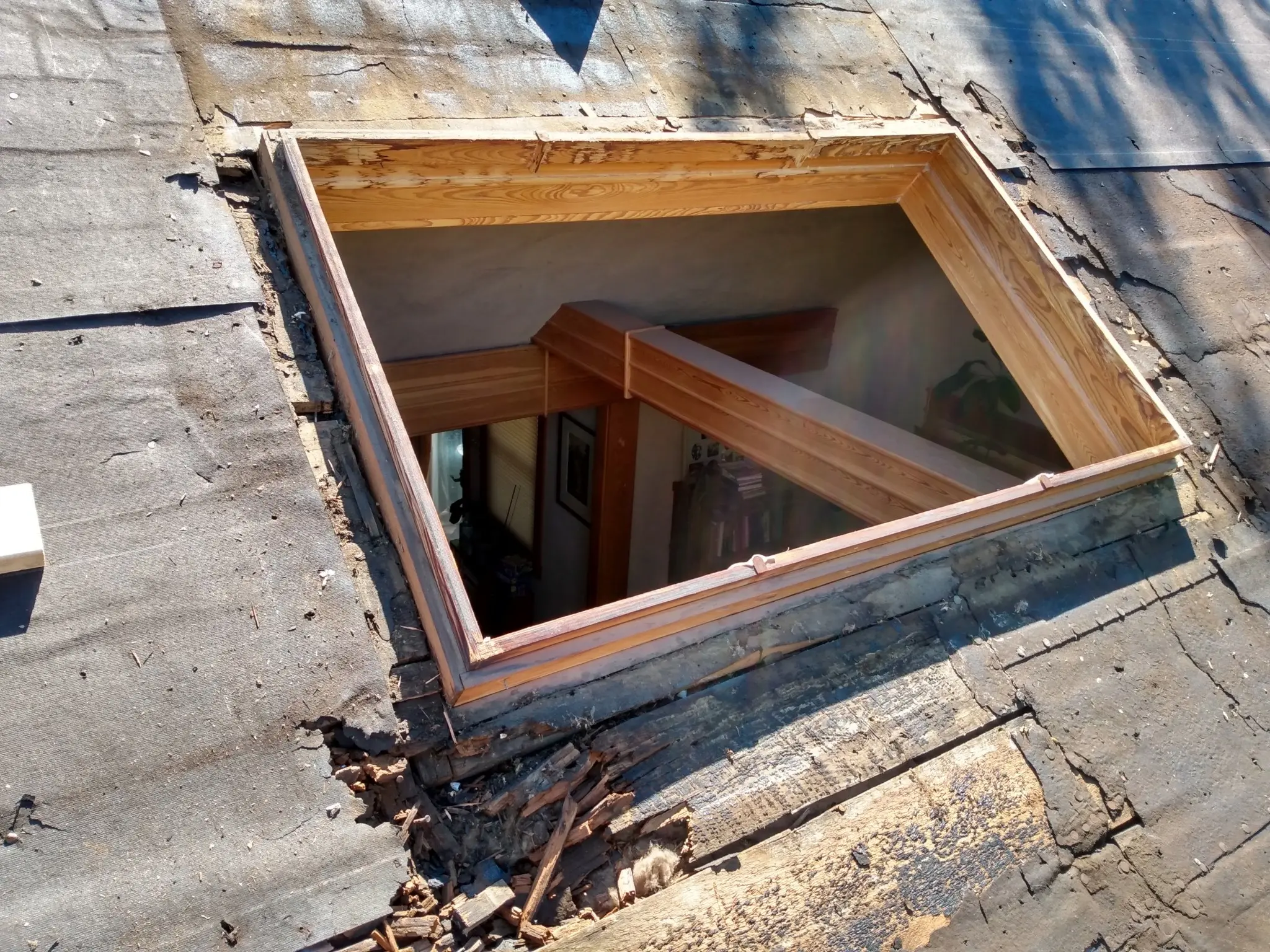 A large rectangular hole with wooden framing has been cut into an old, weathered roof, revealing the interior beams and part of a room inside the house below. Damaged shingles and wood are visible around the opening.