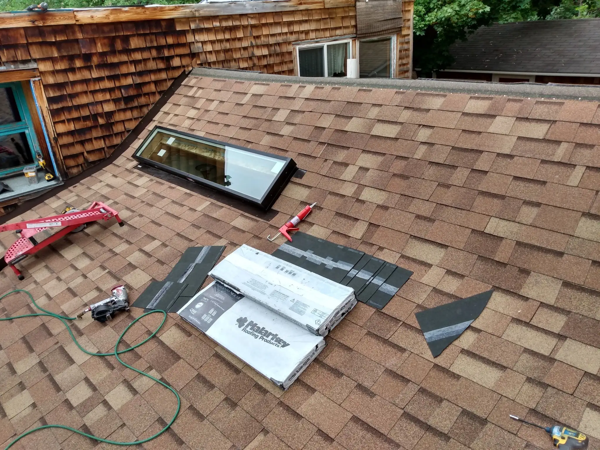 A sloped roof with brown asphalt shingles, a skylight, roofing tools, black shingles, a nail gun, and roofing materials laid out, indicating ongoing roof repair or installation on a residential home.